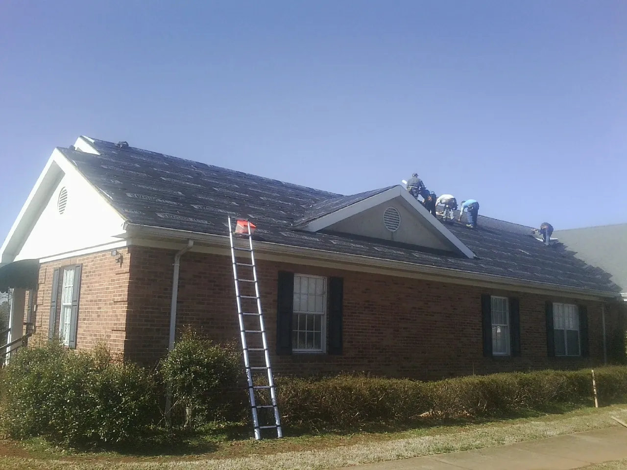 Crew tearing shingles off a brick ranch roof with a ladder set — Emergency Roof Repair work by our crew in Parmele