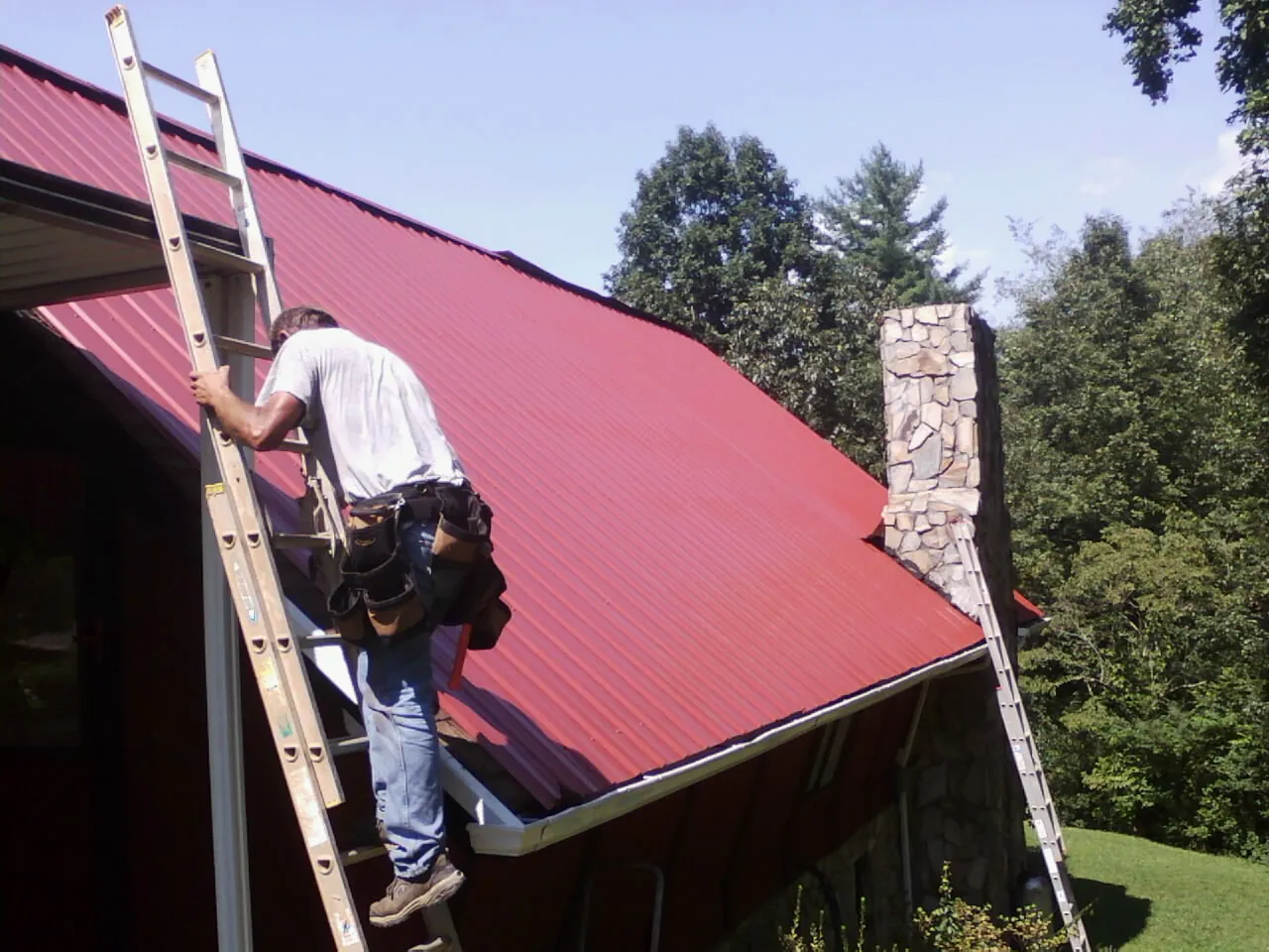 Roofer on a ladder beside a red metal roof and stone chimney — Soffit & Fascia Repair by Roofing Brothers in Parmele